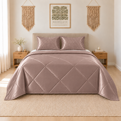 A quilted bedding set in a dusty mauve-pink color, featuring a diamond-quilted comforter and two matching pillow shams, styled on a bed in a warm, neutral-toned bedroom.